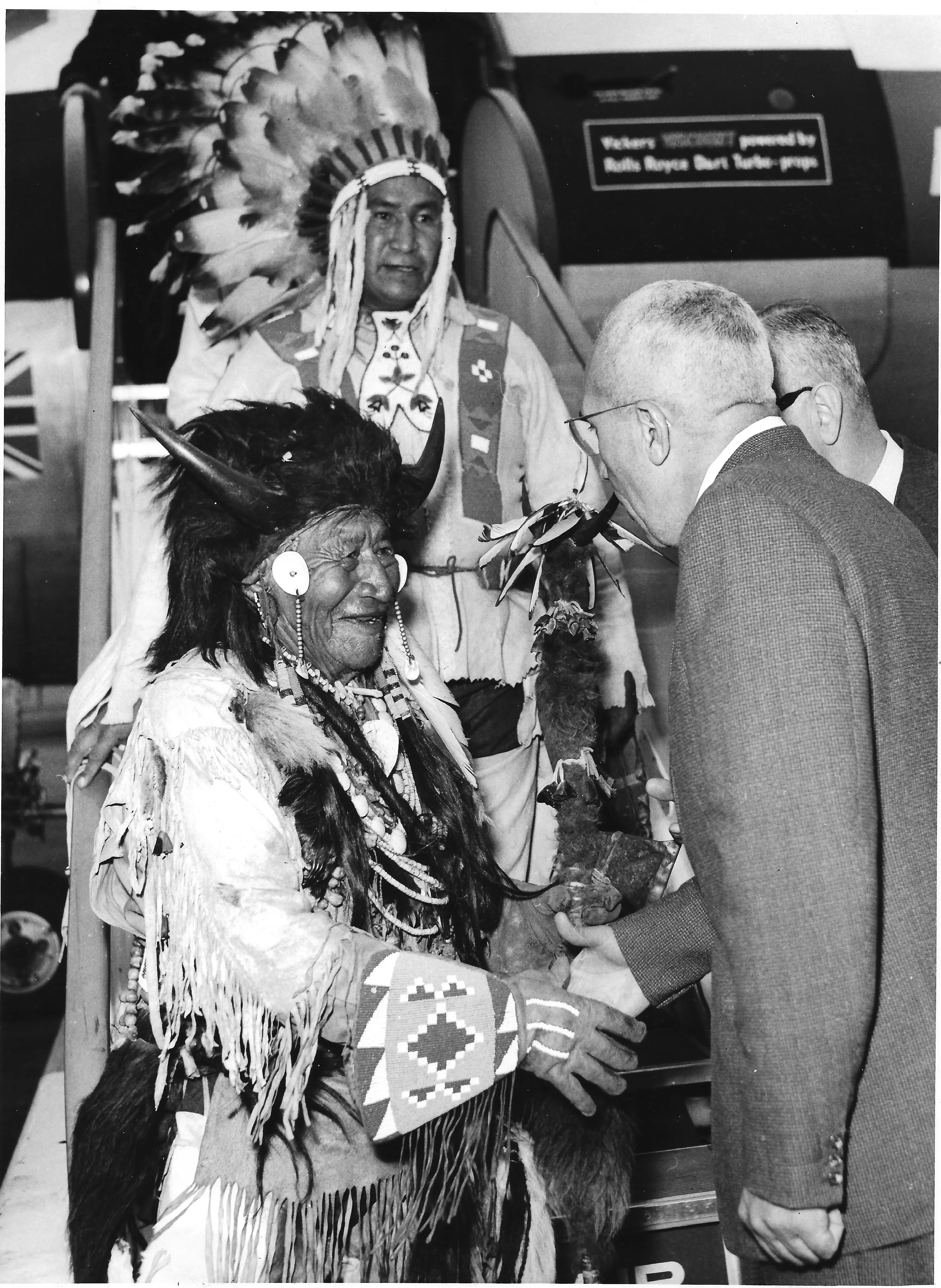 An Indigenous elder in a buffalo-horn headdress and beaded regalia greeting a man in a suit at the foot of an aircraft stairway, a second chief in a feathered headdress behind them, black and white photograph