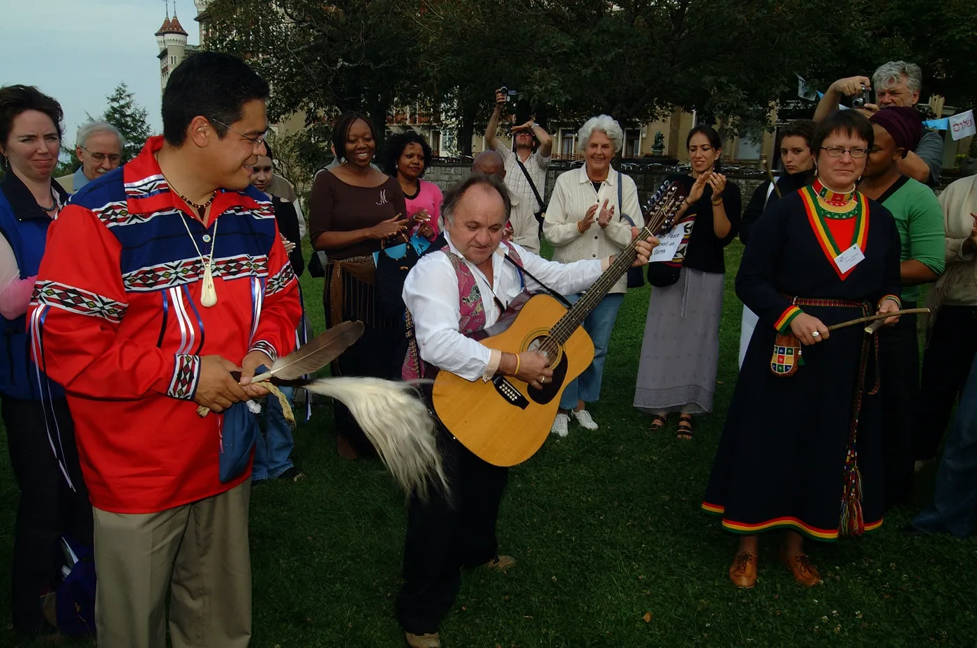 People gathered outdoors in a circle on a lawn with a hand drum and a guitar at a Global Indigenous Dialogue event