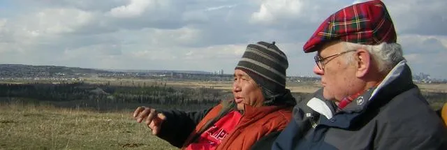 Chief Lee Crowchild seated outdoors on a hillside beside a companion, looking out over the prairie towards a city skyline