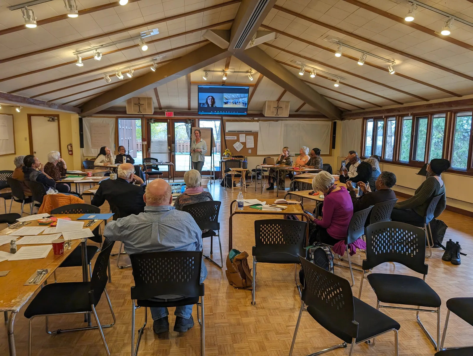 An IofC Canada gathering in a community hall, people seated at tables under a wooden ceiling with tall windows